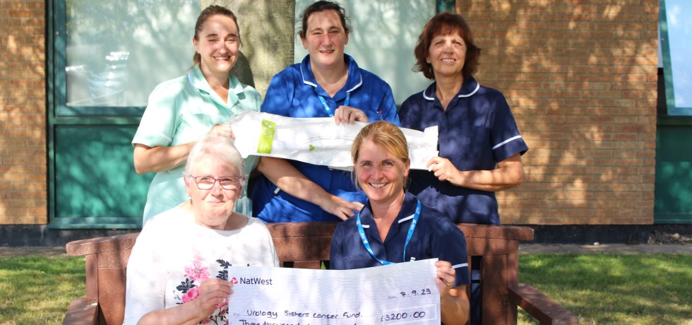 Gloria Joiner and Clare Harvey sitting on a bench holding the giant cheque for £3,200, with other QEH colleagues standing behind.