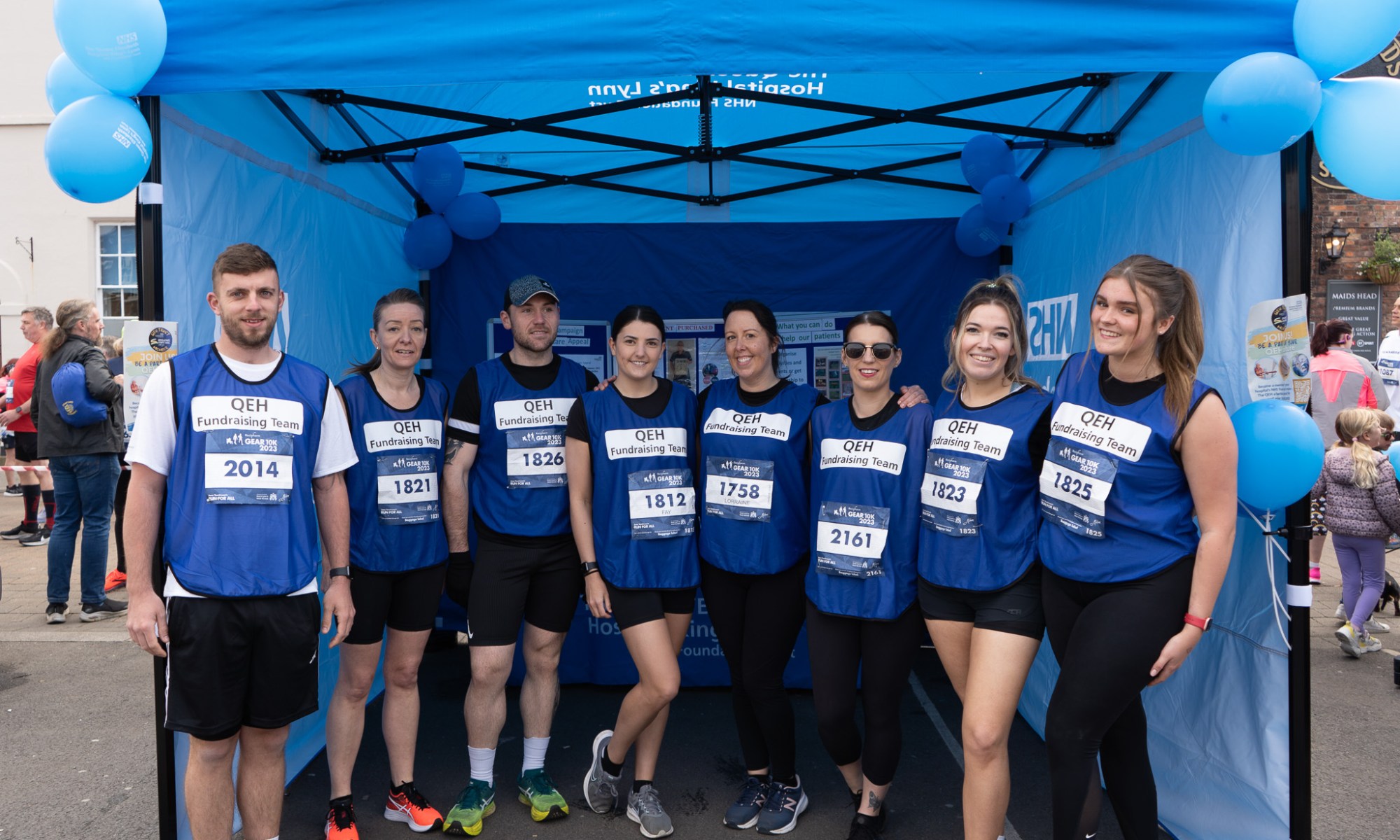 From left to right Jake Kenyon, Emma Southwell, Ryan Oakes, Hannah Auker, Lorraine Hunt, Fay Collison, Hollie Nelson and Ruby Kirby are standing in front of the Queen Elizabeth Hospital gazebo, smiling before the 2023 GEAR 10K race.