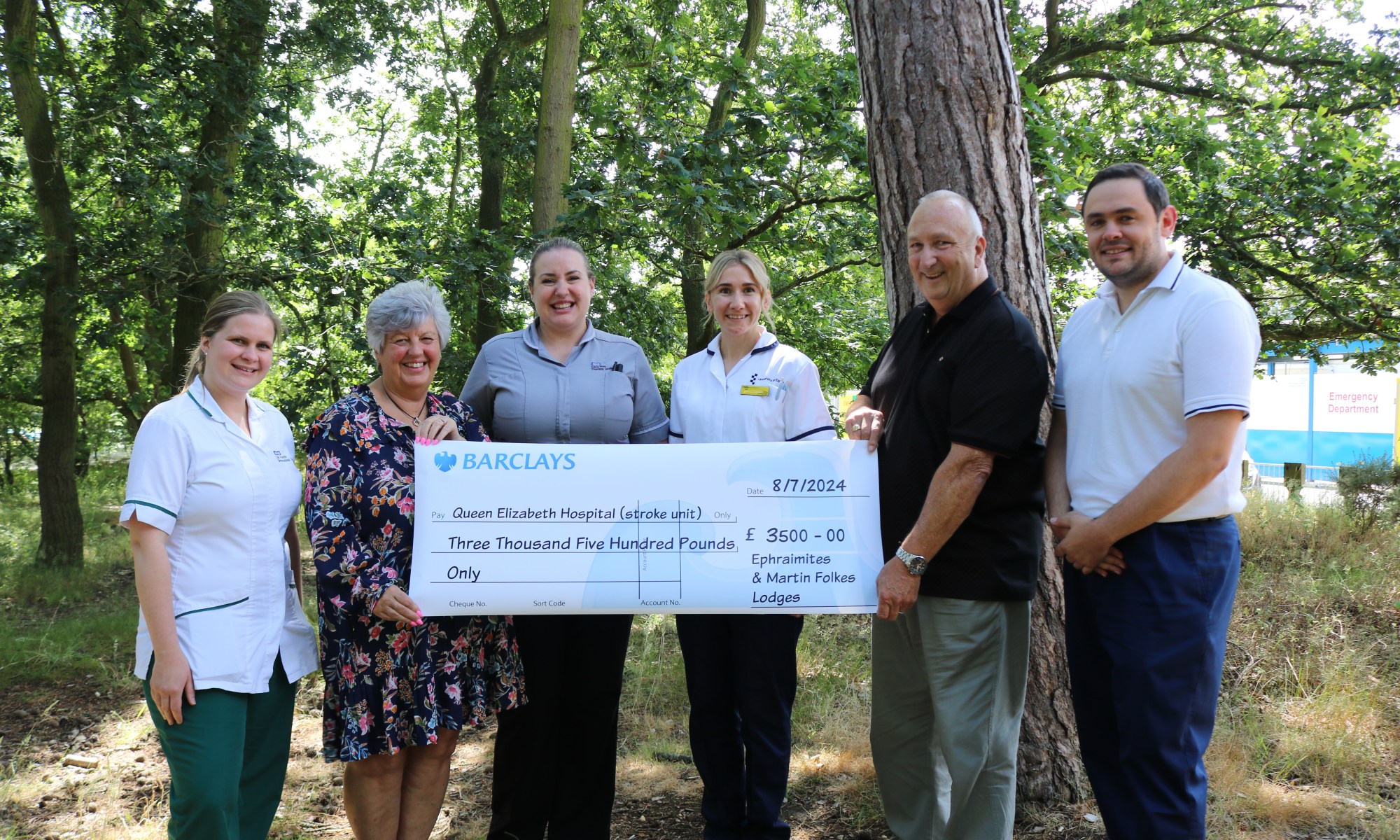 A picture of Mr & Mrs Neill gifting a giant cheque to members of the Queen Elizabeth Hospital stroke team in a woodland area,