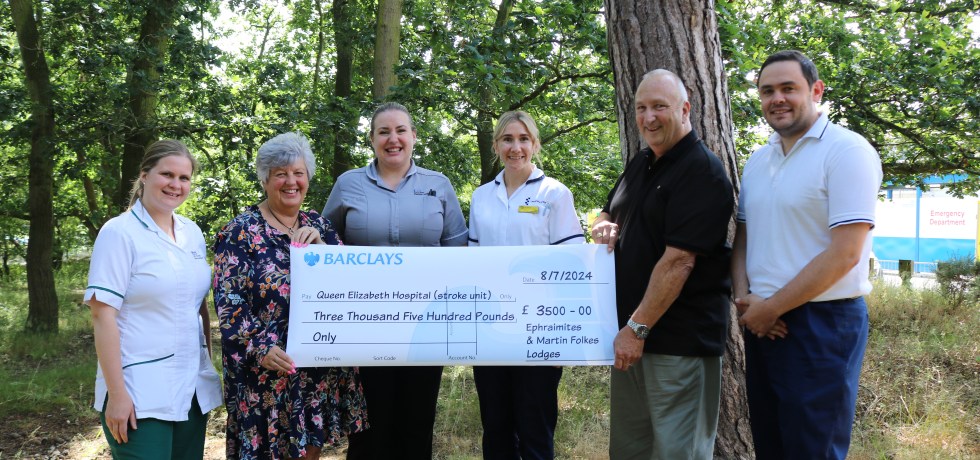 A picture of Mr & Mrs Neill gifting a giant cheque to members of the Queen Elizabeth Hospital stroke team in a woodland area,