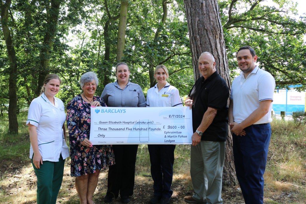 A picture of Mr & Mrs Neill gifting a giant cheque to members of the Queen Elizabeth Hospital stroke team in a woodland area.