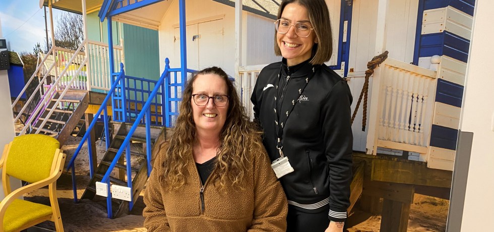 Woman sitting on an exercise ball holding dumbbells, with a personal trainer beside her.