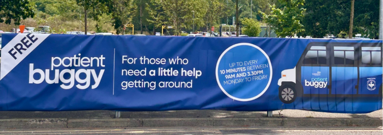 A landscape photo of a fabric banner on railing outside the hospital. The banner depicts the patient buggy. It reads "Free patient buggy. For those who need a little help getting around. Up to every 10 minutes between 9am and 3.30pm Monday to Friday"