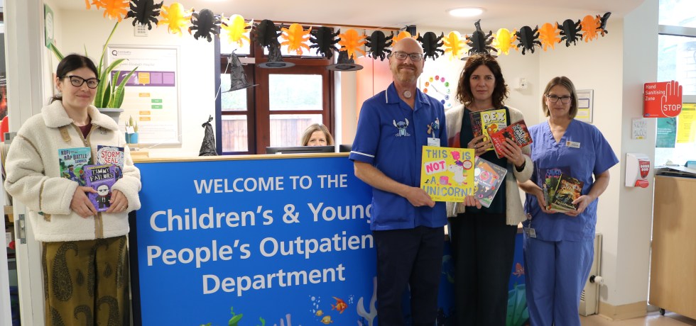 A photo of publishers members with nurses, all holding books standing next to a desk which reads "WELCOME TO THE Children's & Young People's Outpatient Department"