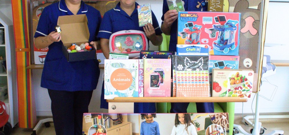 A photo of three nurses holding up various toys in front of a huge pile of toys, activities, play tables, colouring books and video games.