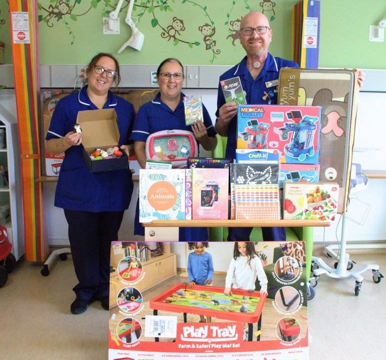 A photo of three nurses holding up various toys in front of a huge pile of toys, activities, play tables, colouring books and video games.