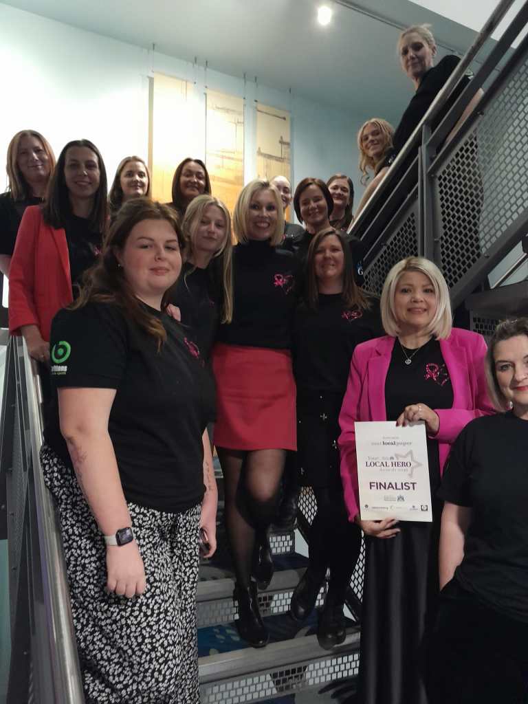 Members of Breast of Friends holding up a "Your Local Hero Awards 2026 Finalist" certificate on a staircase.