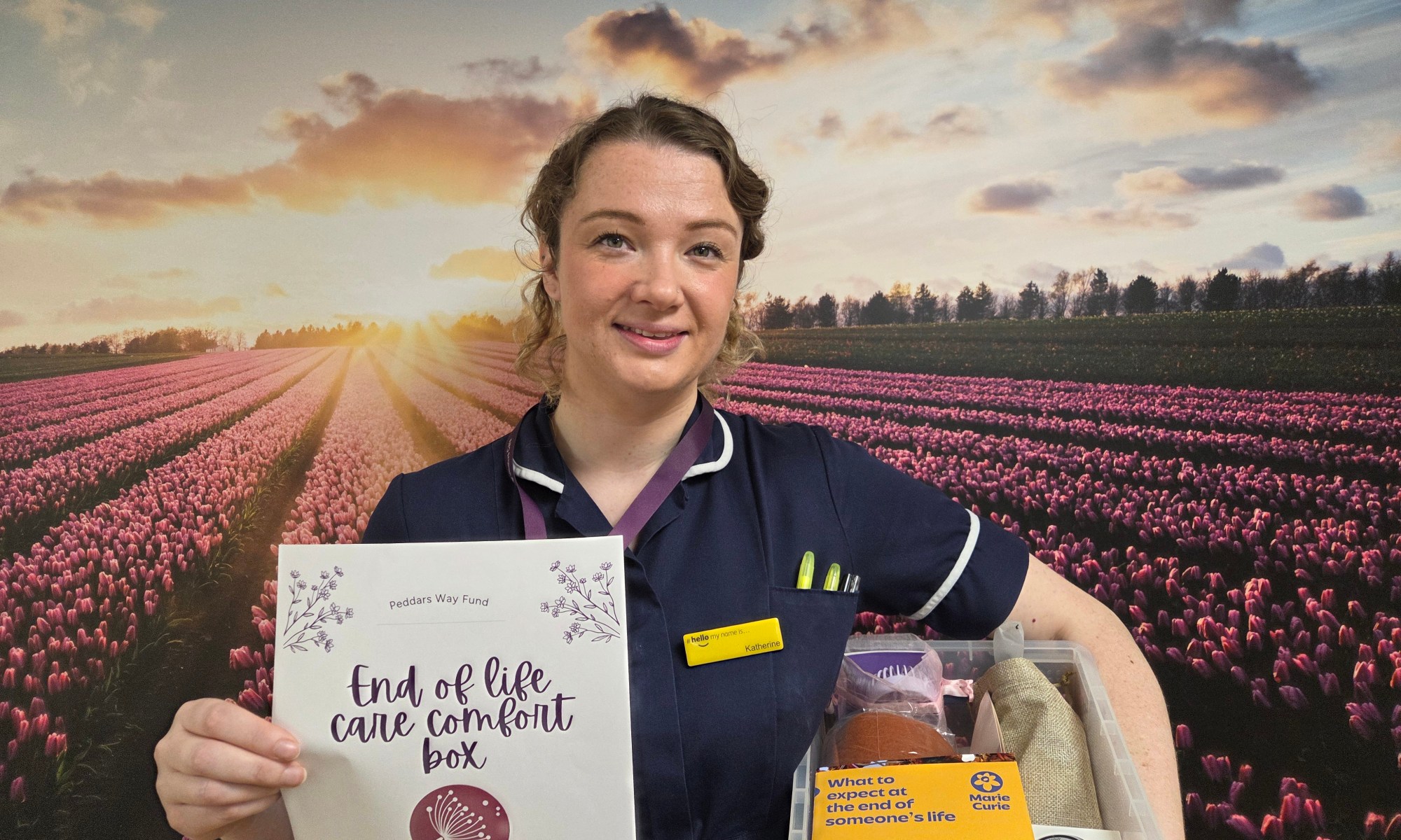 A nurse with blond hair holding a box and a poster in front of a purple background