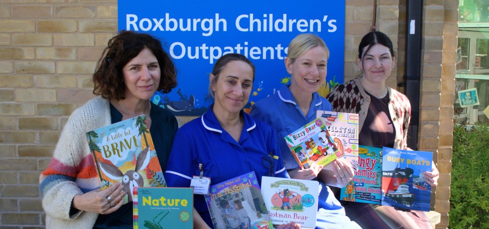 A photo of two publisher representatives sitting next to two nurses outside holding children's books. The sign behind them reads "Welcome to the Roxburgh Children's Outpatients"