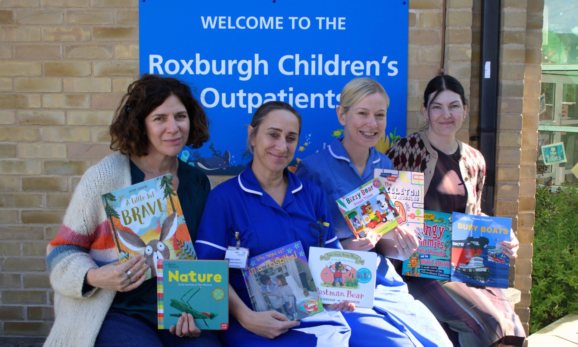 A photo of two publisher representatives sitting next to two nurses outside holding children's books. The sign behind them reads "Welcome to the Roxburgh Children's Outpatients"