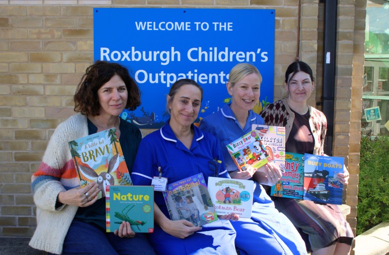 A photo of two publisher representatives sitting next to two nurses outside holding children's books. The sign behind them reads "Welcome to the Roxburgh Children's Outpatients"
