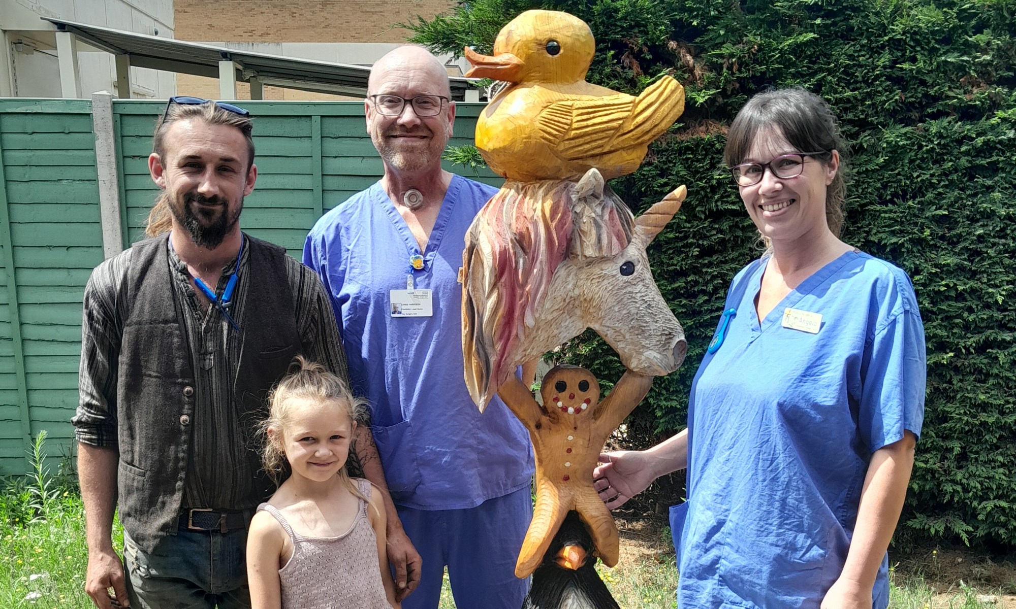 A photo of two nurses stood next to Billy Bicknell and Jeana next to a large totem pole consisting of a capybara, a penguin, a gingerbread man, a unicorn and a duck.
