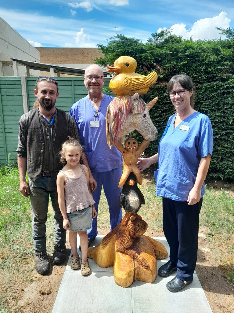 A photo of two nurses stood next to Billy Bicknell and Jeana next to a large totem pole consisting of a capybara, a penguin, a gingerbread man, a unicorn and a duck.