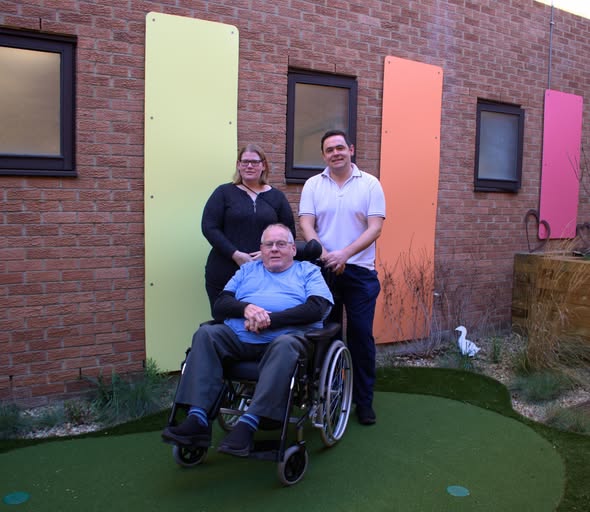 A photo of Maggie Drew with a stroke rehabilitation nurse and Maggie's farther in the specialised wheelchair in the QEH's Stroke Rehabilitation Garden