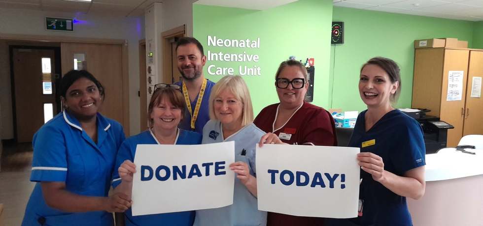 A photo of NICU nurses & clinicians holding up two sheets of paper that read "Donate Today!" written on them. The ward shows "Neonatal Intensive Care Unit" on the green wall in the background.