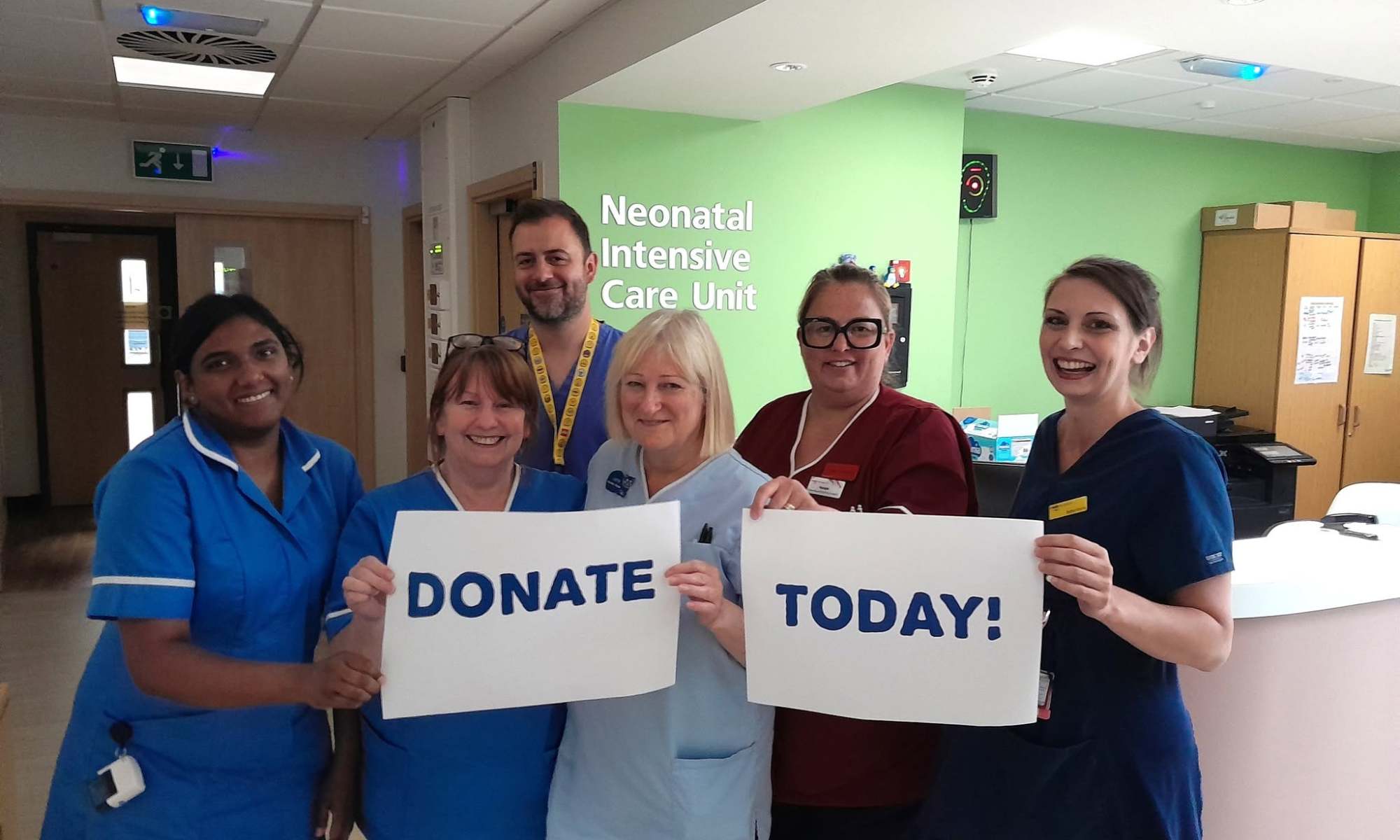 A photo of NICU nurses & clinicians holding up two sheets of paper that read "Donate Today!" written on them. The ward shows "Neonatal Intensive Care Unit" on the green wall in the background.