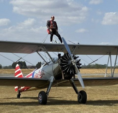 A photo of Brian Moss strapped to the top of an airplane. On the side the airplane, the text "The Wing Walk Company" can be seen