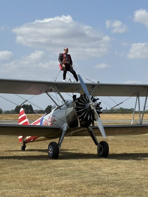 A photo of Brian Moss strapped to the top of an airplane. On the side the airplane, the text "The Wing Walk Company" can be seen