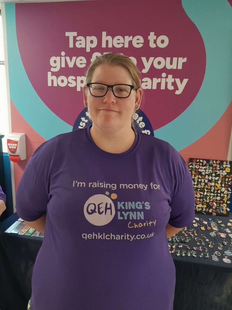 A photo of Maggie Drew smiling in front of a stall wearing a "I'm raising money for The QEHKL Charity" t shirt