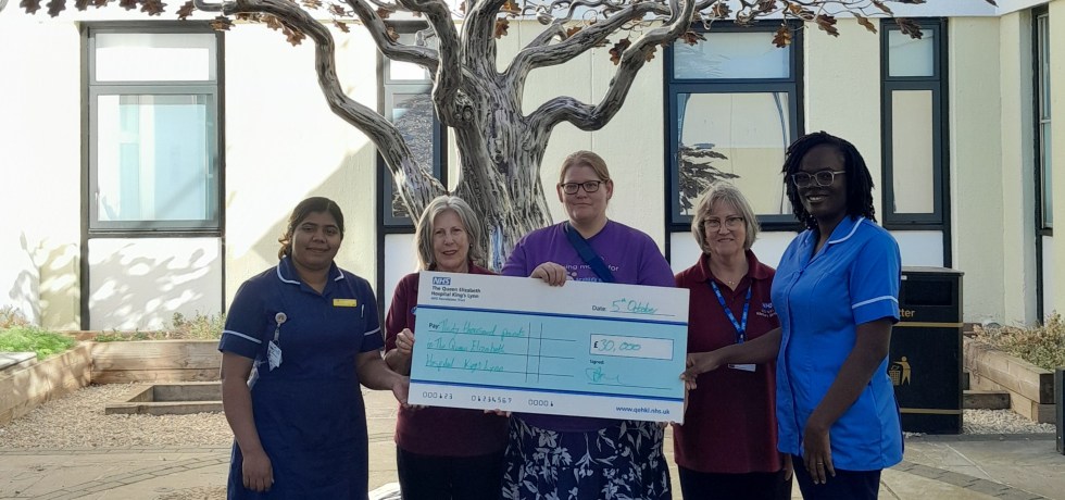 A photo of Maggie Drew holding a giant novelty cheque worth £30,000 to The Queen Elizabeth Hospital Charitable Fund, next to Cancer Ward Manager, a nurse and two volunteers.