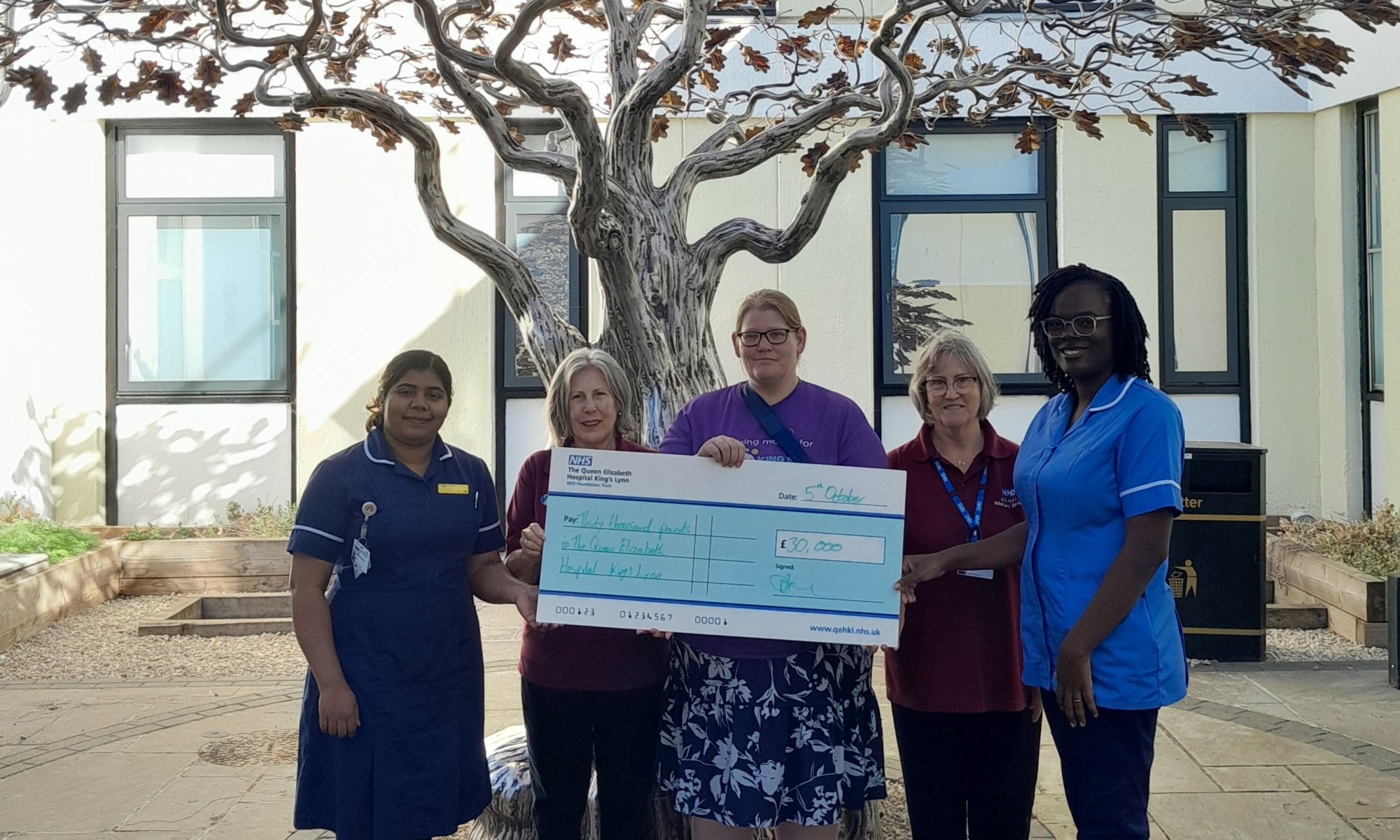 A photo of Maggie Drew holding a giant novelty cheque worth £30,000 to The Queen Elizabeth Hospital Charitable Fund, next to Cancer Ward Manager, a nurse and two volunteers.