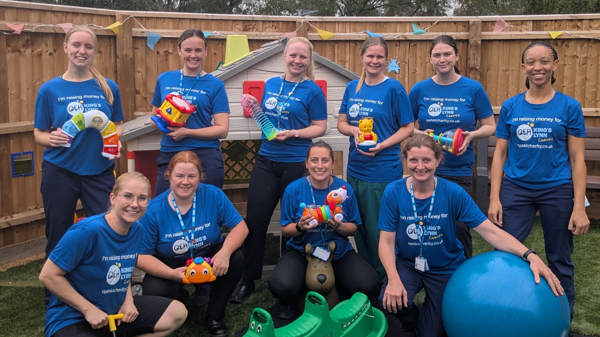 A photo of the play team wearing "I'm raising money for QEHKL Charity" holding various toys in an outdoor play area.